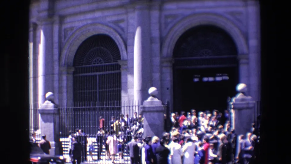 1967 Madrid home-movie frame showing pedestrians gathered near a wrought iron fence and arched stone building entrance, with strong sunlight and shadow.