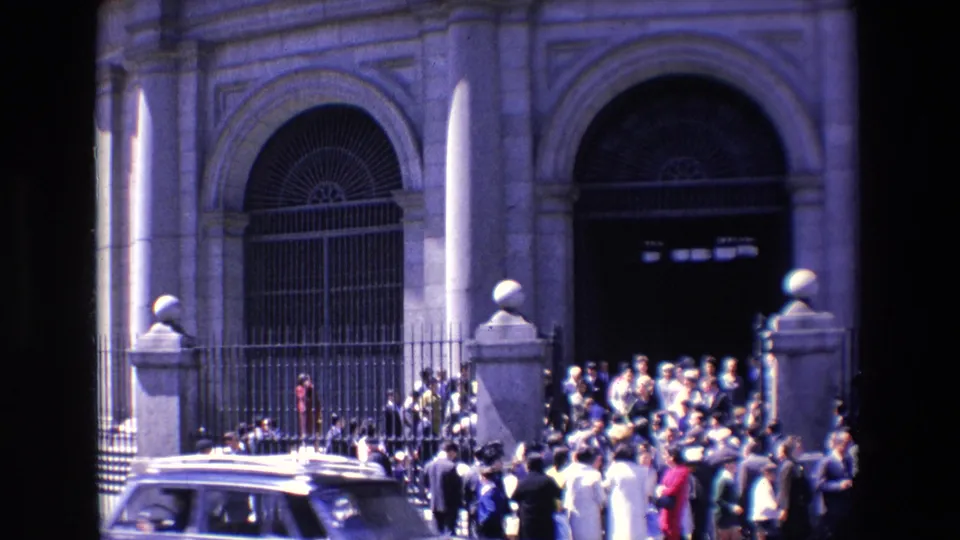 1967 Madrid street view with a car or van crossing the foreground while people cluster near an iron fence and large arched doorway.
