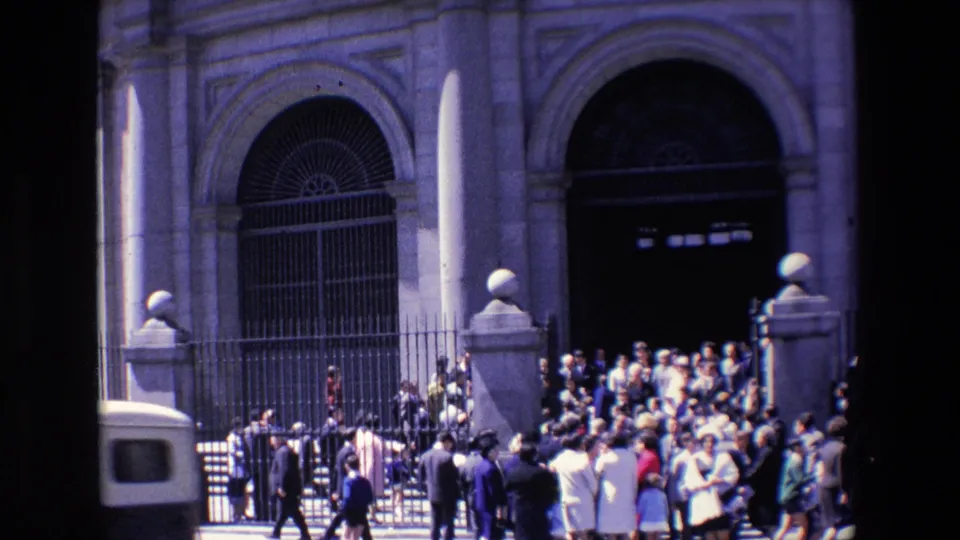 1967 Madrid frame showing pedestrians moving along the street as a light-colored vehicle passes beside a crowd gathered at a fenced stone facade.