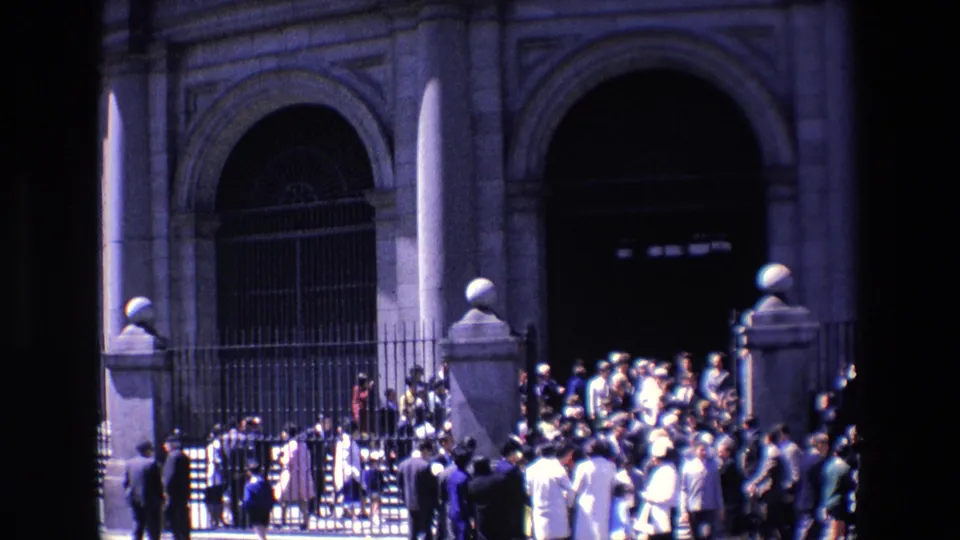 1967 Madrid home-movie frame with a dense crowd near a dark arched entrance, stone pillars, and iron fencing along a sunlit street.
