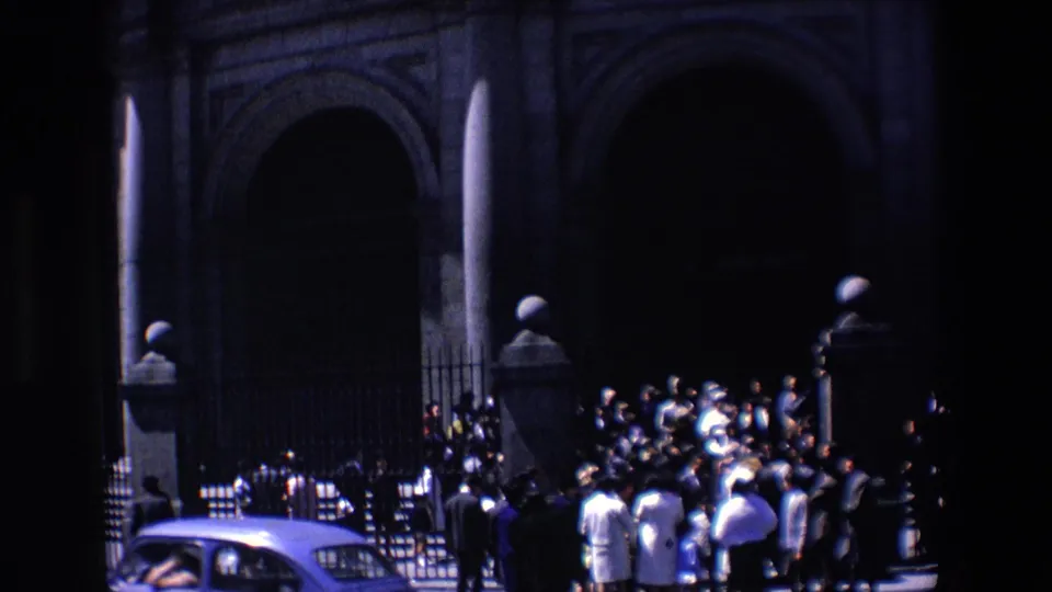 1967 Madrid home-movie frame showing a crowd outside a large arched stone entrance, with a pale car partly visible at the lower left.
