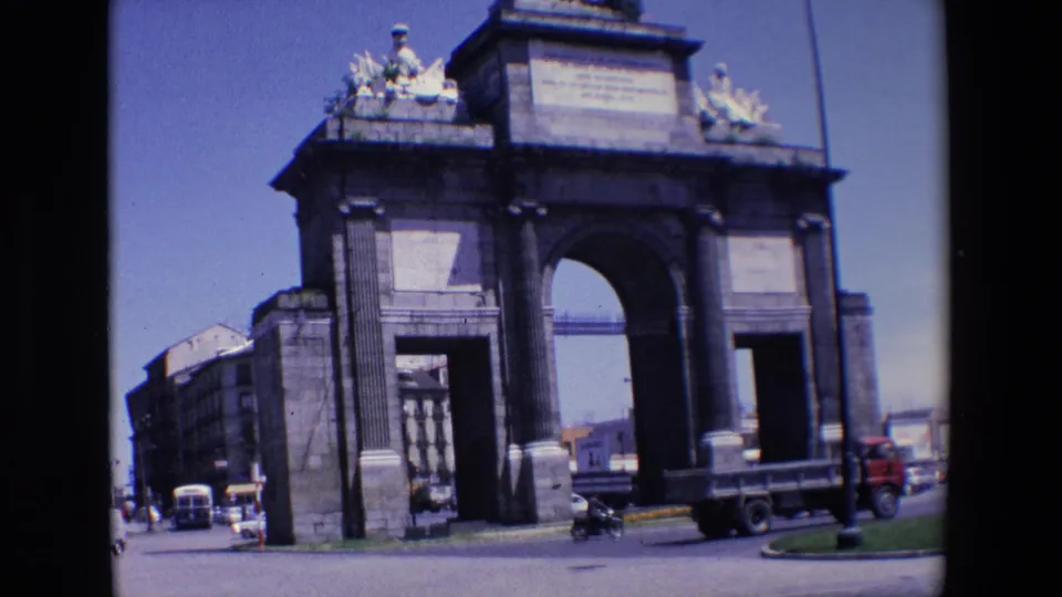 1967 Madrid home-movie frame of a monumental stone gateway from across the street, with cars, a scooter, buildings, and blue sky visible.