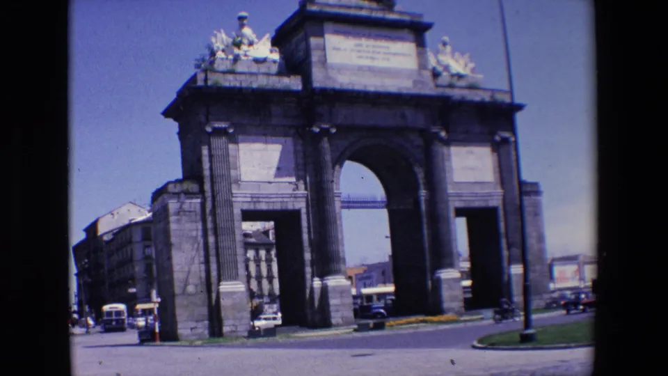 1967 Madrid frame showing the stone monument's arched openings beside a roadway, with cars, a street lamp, and clear blue sky.