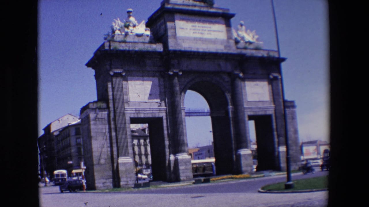 1967 home-movie frame of a large stone triumphal arch in Madrid with cars and a bus moving along the road in front.