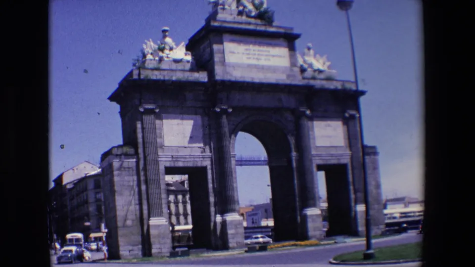 1967 Madrid travel-film view of a monumental stone arch, surrounding buildings, and mid-century vehicles moving through the street scene.