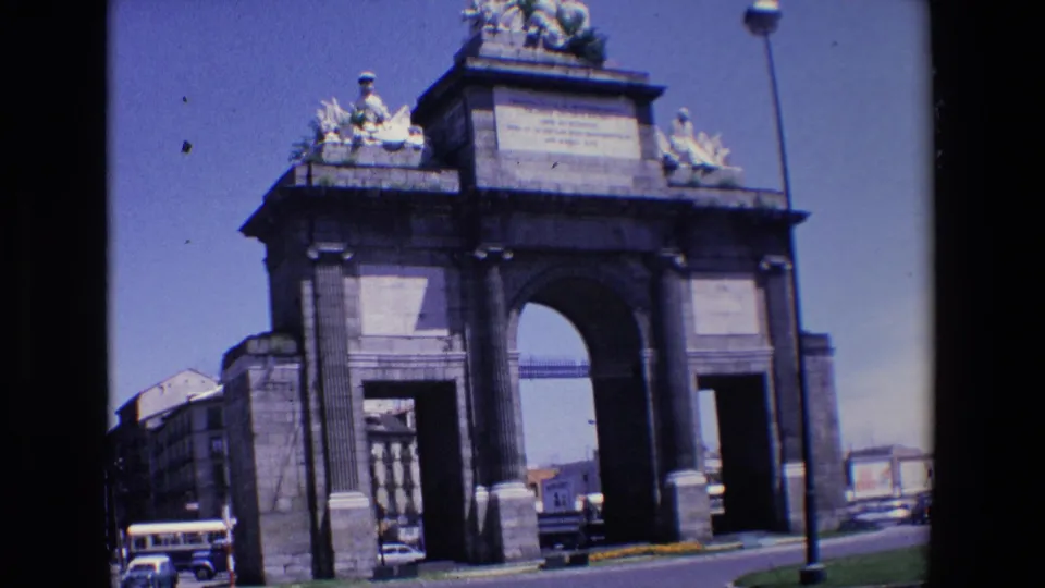 Handheld 8mm frame from Madrid in 1967 showing a stone triumphal arch, a bus at the roadside, and cars moving through the surrounding traffic.