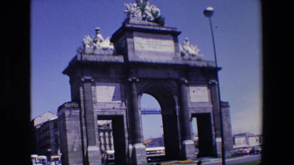1967 Madrid home-movie frame showing the monumental arch from across the road, with vehicles, city buildings, and a tall streetlamp in view.