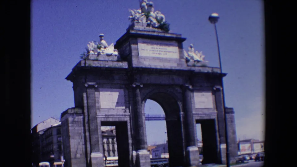 1967 Madrid home-movie frame showing a monumental arched stone gateway with sculptural figures above, inscription panels, a streetlamp, and cars beyond the opening.