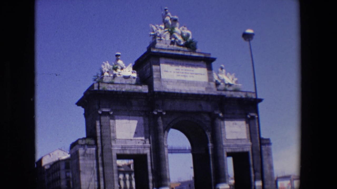 Arched monument with sculptural roofline, side panels, streetlamp, and distant buildings.
