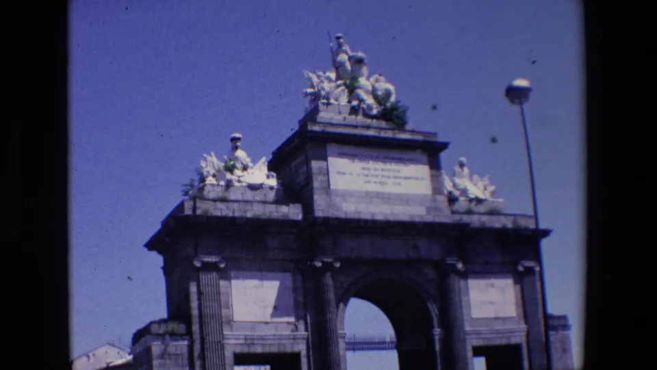1967 Madrid home-movie frame with a slightly tilted closer view of the gateway, emphasizing the rooftop sculpture group, column details, inscription panel, and arched opening.