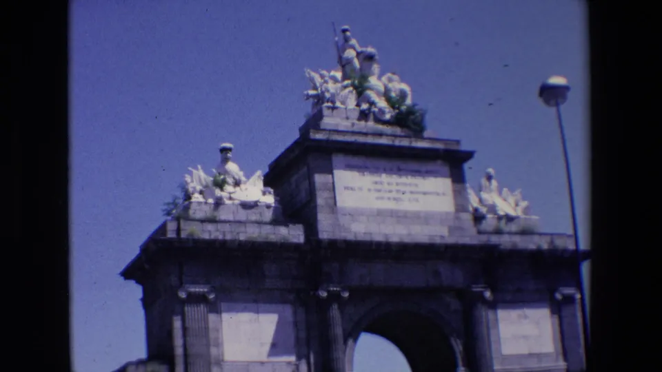 1967 Madrid frame showing the monument from a low street-level angle, with the carved roofline, central inscription block, dark arch opening, side stonework, and streetlamp visible.