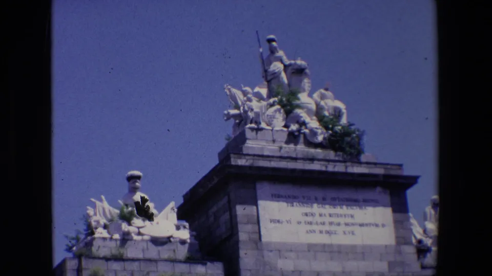 1967 Madrid home-movie frame with a closer upward view of the monument's sculptural crown, inscription block, and weathered stonework.