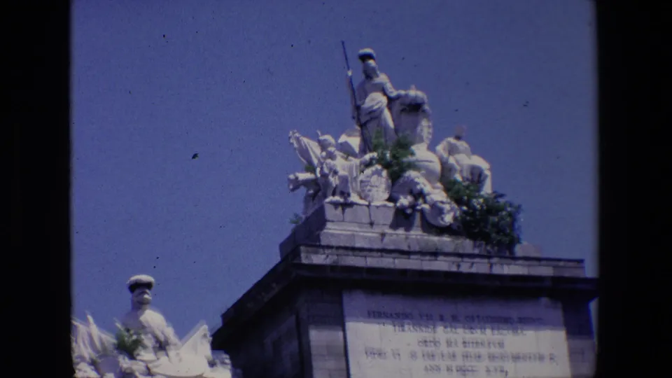 Final 1967 Madrid frame showing the upper monument from a low angle, including sculptural figures, foliage, and a partially legible inscription panel.