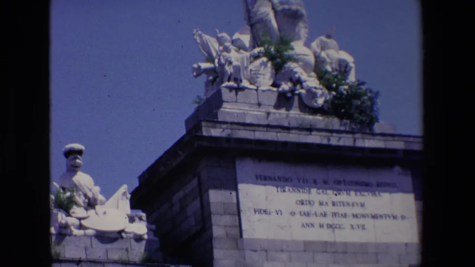 1967 Madrid 8mm frame showing a stone commemorative structure with a carved inscription panel, partial sculptural figures above the cornice, and a dark film edge along the left side.
