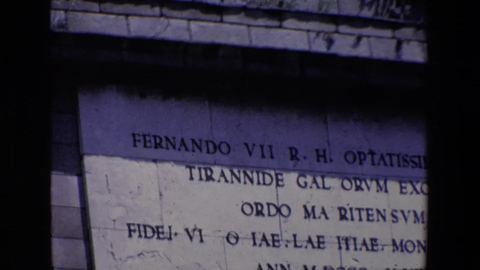 1967 Madrid 8mm frame showing a cropped carved inscription on a stone monument, with the upper line partly legible and a heavy dark film-edge shadow through the center.