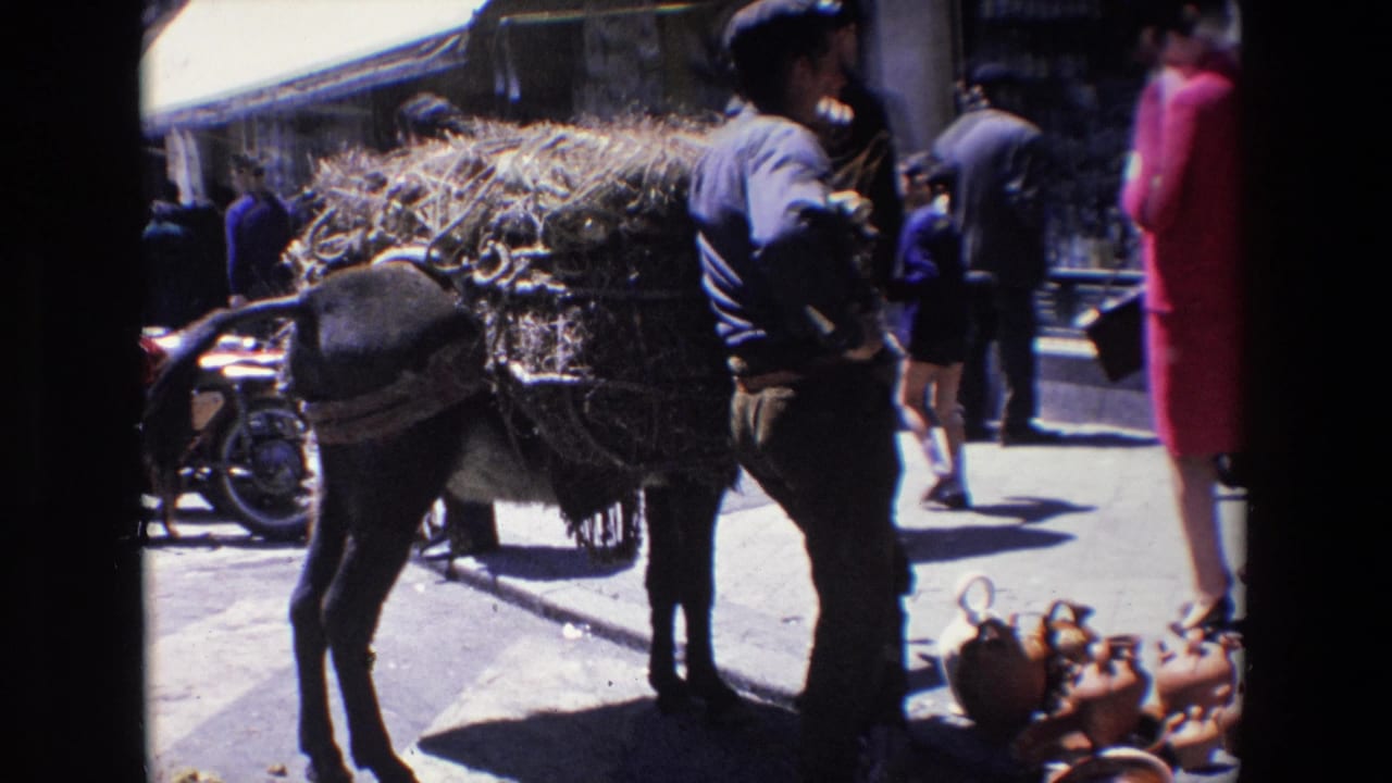 Donkey loaded with hay beside a man, with pedestrians and pottery on the pavement.