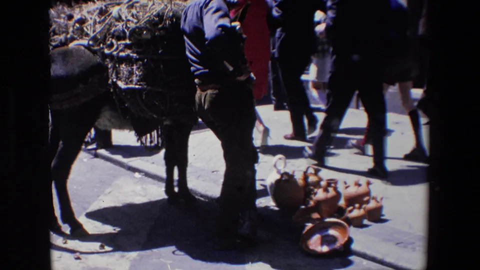 1967 Madrid home-movie frame showing the donkey and handler near a pavement display of clay vessels as pedestrians pass through the street.