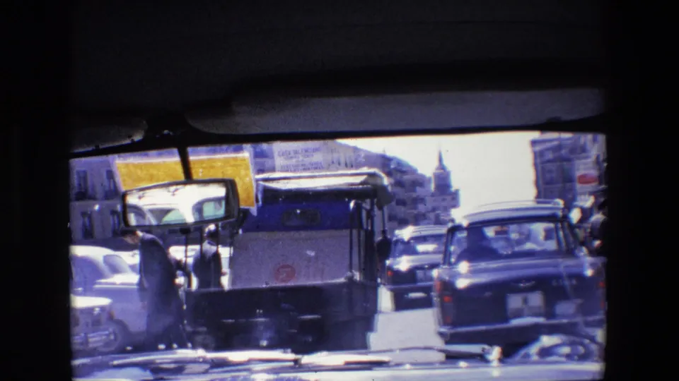1967 Madrid traffic scene seen through a dark window frame, with compact cars, a small utility vehicle, and pedestrians along a bright urban street.