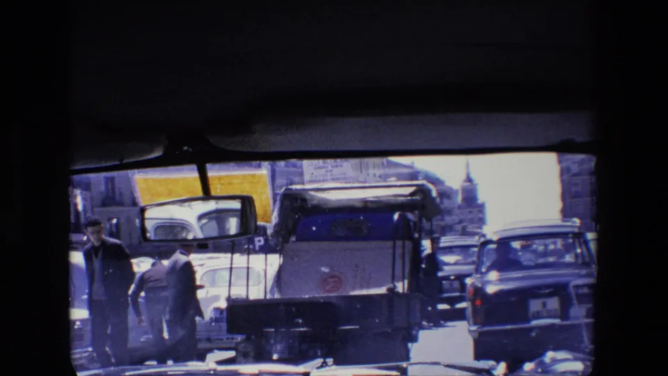 1967 Madrid home-movie frame with pedestrians crossing or standing near compact cars and a small utility vehicle in bright urban traffic.
