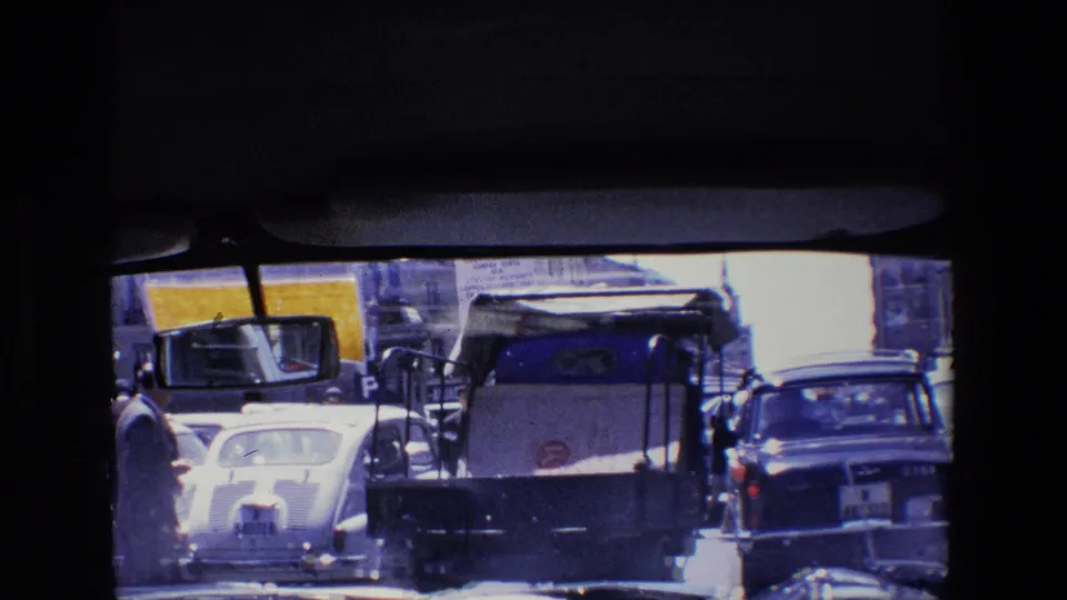 1967 Madrid traffic frame showing the backs of compact cars, a small utility vehicle, pedestrians at the street edge, and dark home-movie frame borders.