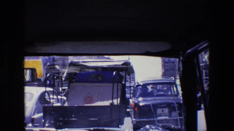 1967 Madrid street traffic viewed from inside a vehicle, with a white service vehicle, cars, windshield framing, and buildings beyond.