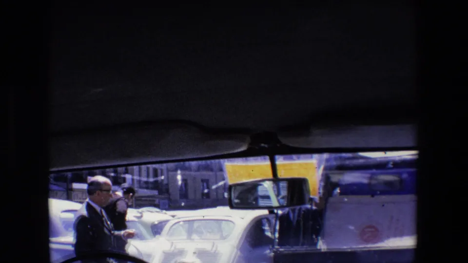Pedestrians in formal clothing wait or move beside cars and a white service vehicle on a congested 1967 Madrid street.