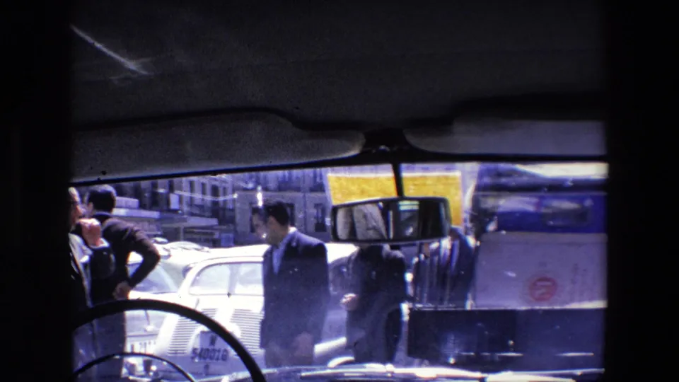 Men in suits stand near cars and a service vehicle in 1967 Madrid traffic, framed by the dark interior of the filming vehicle.