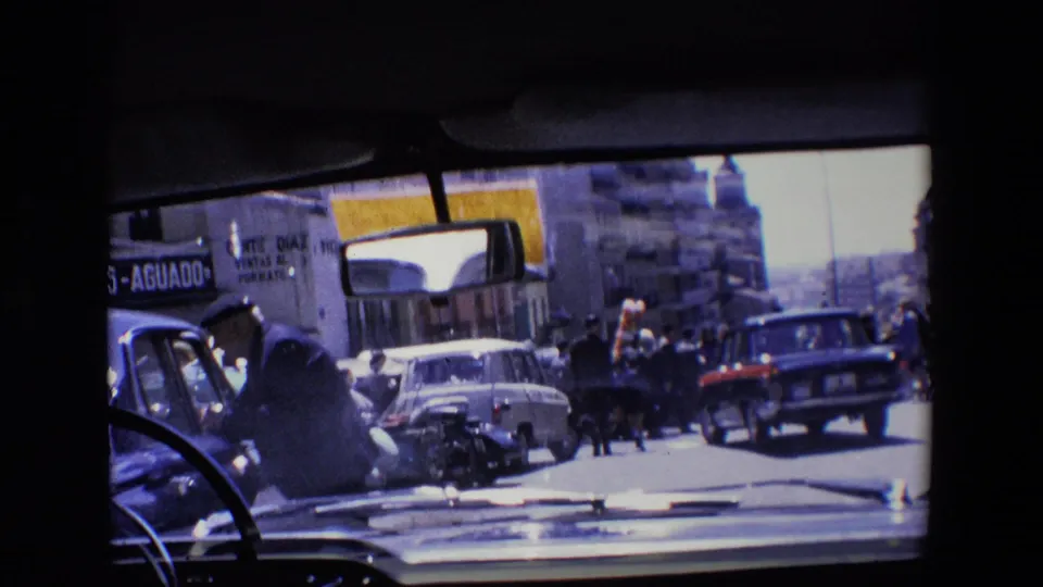 A congested 1967 Madrid street shows cars, pedestrians, storefront signage, and dashboard framing from inside the filming vehicle.