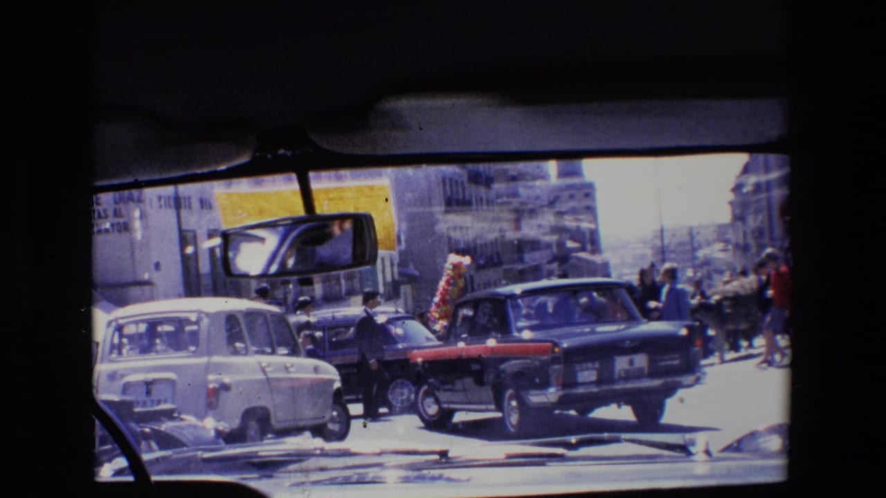Cars pass along a busy street while pedestrians gather near buildings.