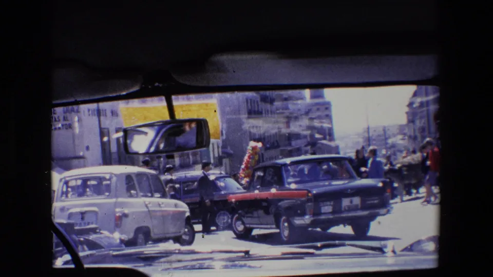 1967 Madrid street view from inside a vehicle, showing a dark sedan, a white car, pedestrians, building facades, and a rear-view mirror in the foreground.