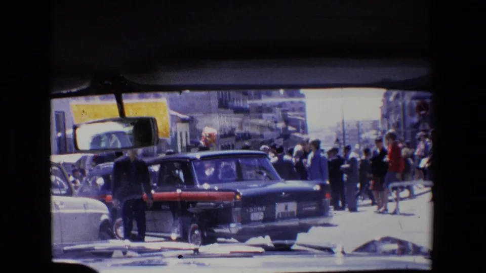 1967 Madrid home-movie frame with a dark car close to the camera, pedestrians along the sidewalk, windshield edges, and sunlit urban buildings in the background.