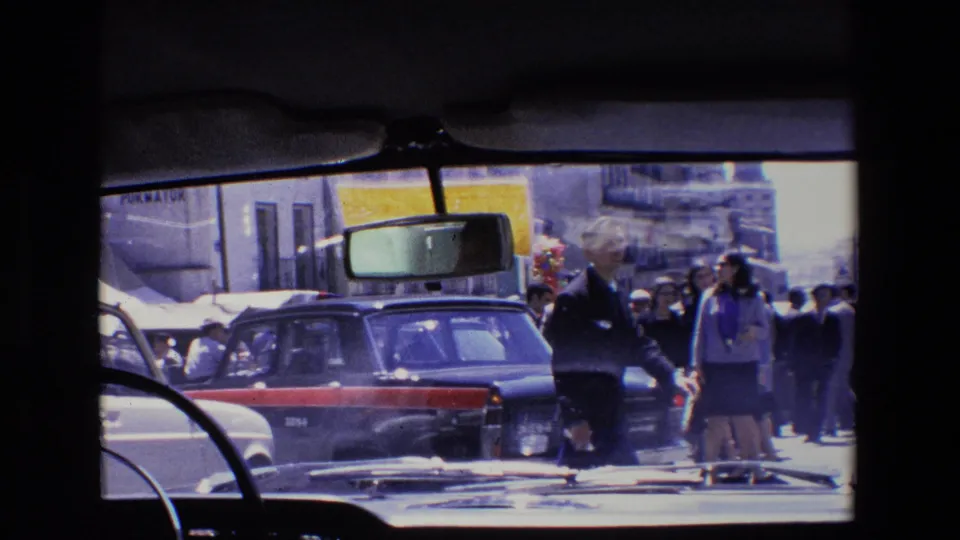 1967 Madrid street scene filmed from inside a vehicle, with a dark car in the foreground, people walking or standing along the curb, building fronts, and windshield details visible.