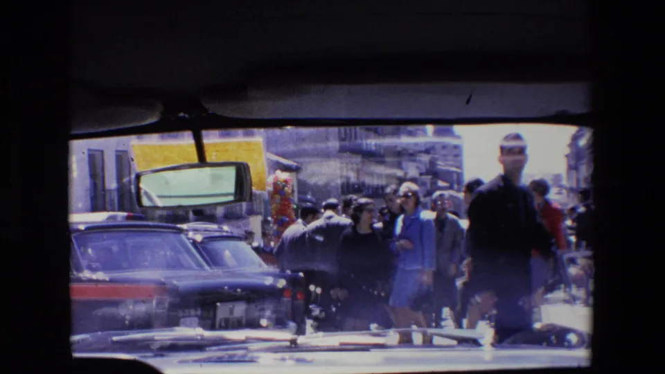 1967 Madrid street view with pedestrians walking past older cars and storefronts, filmed through dark window-like framing.