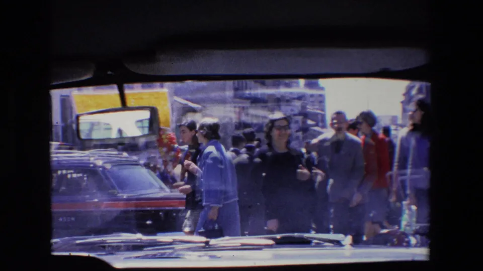 Crowded 1967 Madrid sidewalk where men and women pass cars and shopfronts in bright daylight.