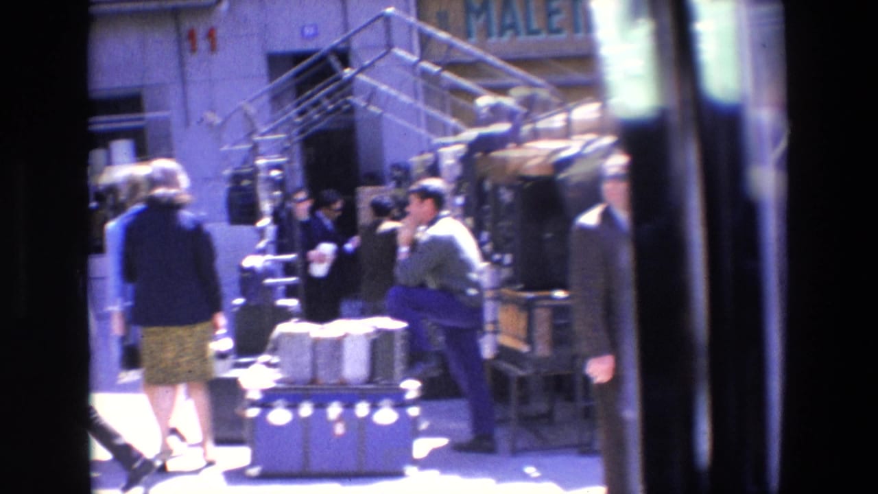 Pedestrians pass outdoor display tables and a shopfront on a sunny street.
