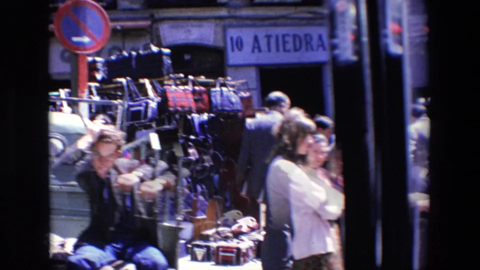 Madrid 1967 home-movie frame showing sidewalk racks with bags or garments, a seated person, passing pedestrians, parked cars, and a no-parking sign.