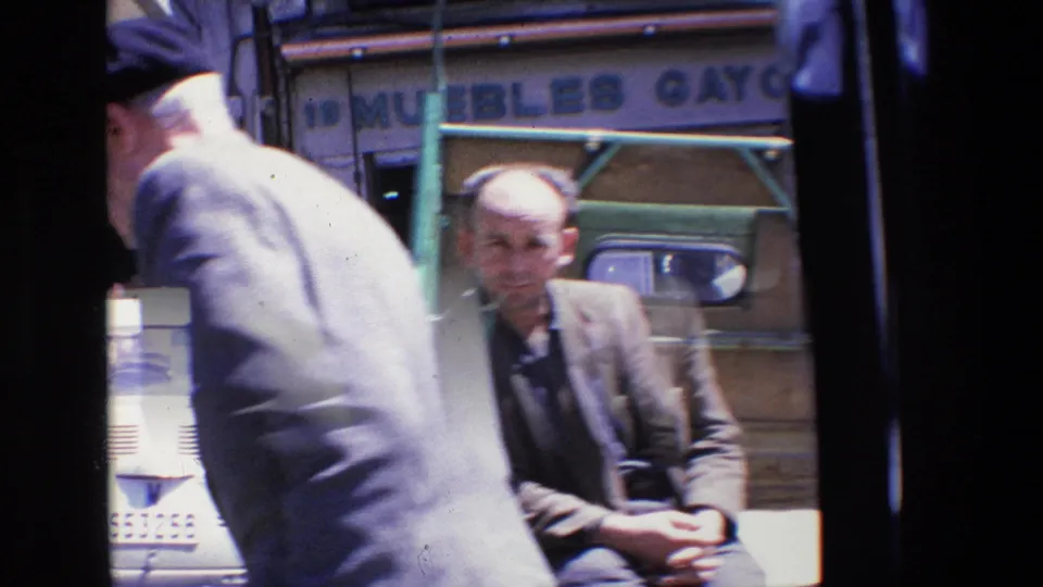 Madrid 1967 street frame with two men in suits near a storefront awning, a parked vehicle, and partial Spanish shop lettering visible behind them.