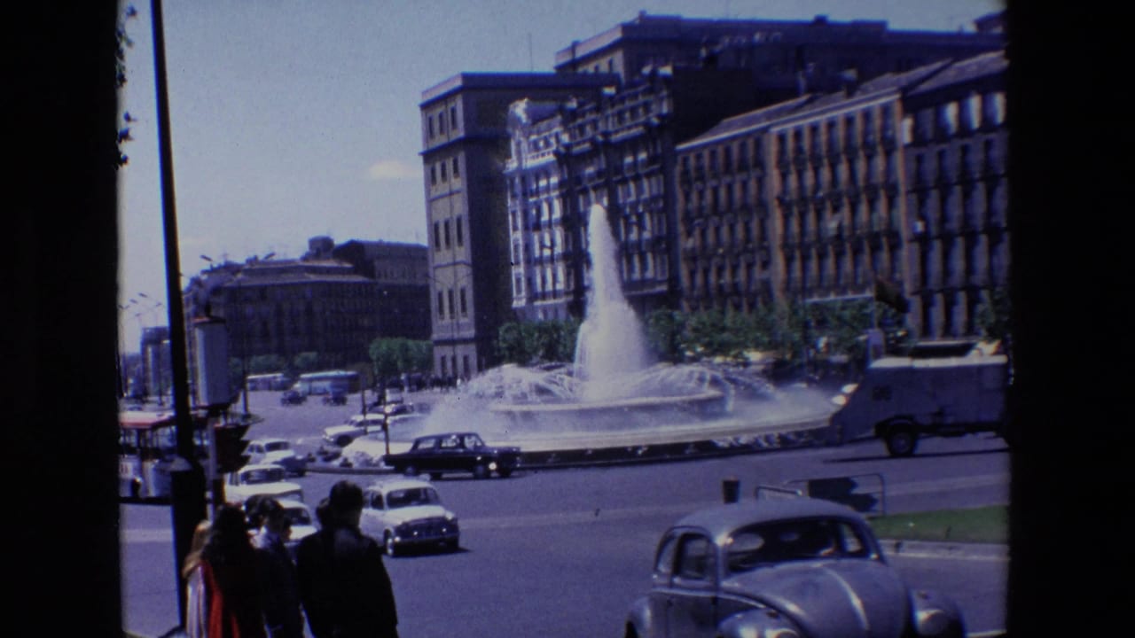 1967 Madrid home-movie view of cars circling a large public fountain, with pedestrians near the camera and tall city buildings behind it.