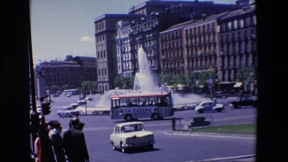1967 Madrid home-movie frame with a small bus bearing visible side advertising, cars, foreground pedestrians, and a large fountain against city buildings.