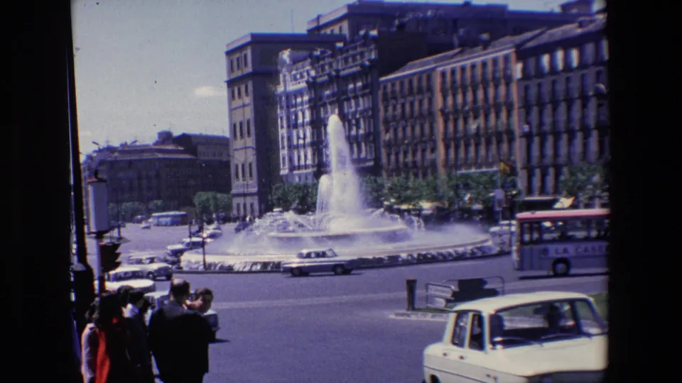 1967 Madrid street footage frame with a light-colored car in the foreground, pedestrians at lower left, and the spouting public fountain behind moving traffic.