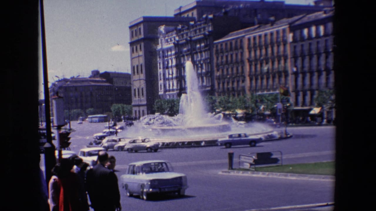 Traffic circle with a large fountain, cars, pedestrians, and tall city buildings.