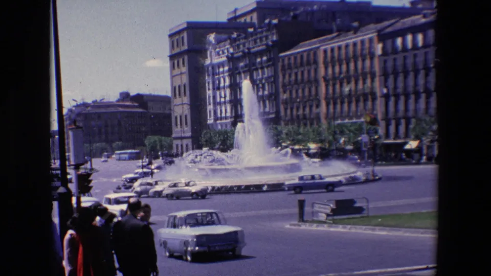 1967 Madrid street view showing a large water fountain in a traffic circle with cars, pedestrians at the curb, and surrounding masonry buildings.
