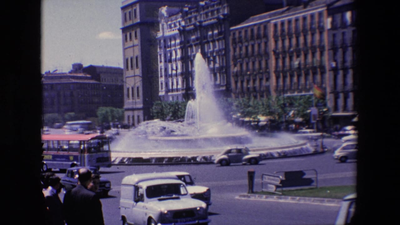 Bus and cars move around a fountain in front of tall urban buildings.