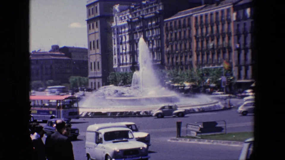 1967 Madrid traffic-circle view with a city bus, cars, pedestrians, a spraying fountain, and tall buildings lining the roadway.