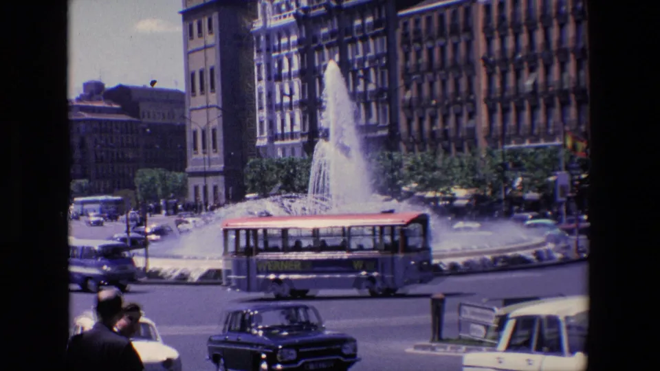 1967 Madrid home-movie frame showing a bus with visible side lettering passing the fountain while cars and pedestrians move through the roundabout.