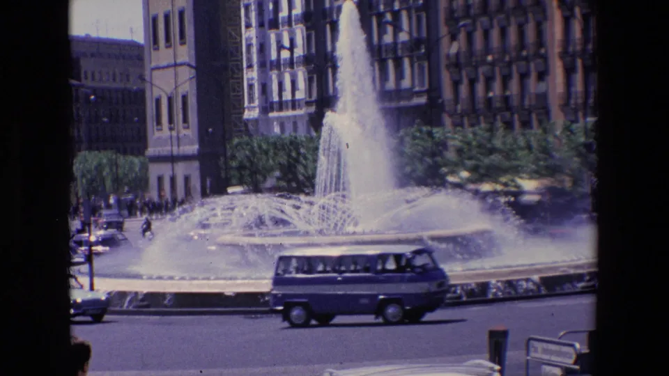1967 Madrid street scene with a van crossing the foreground, cars near the curb, a large fountain, trees, and tall buildings around the circle.