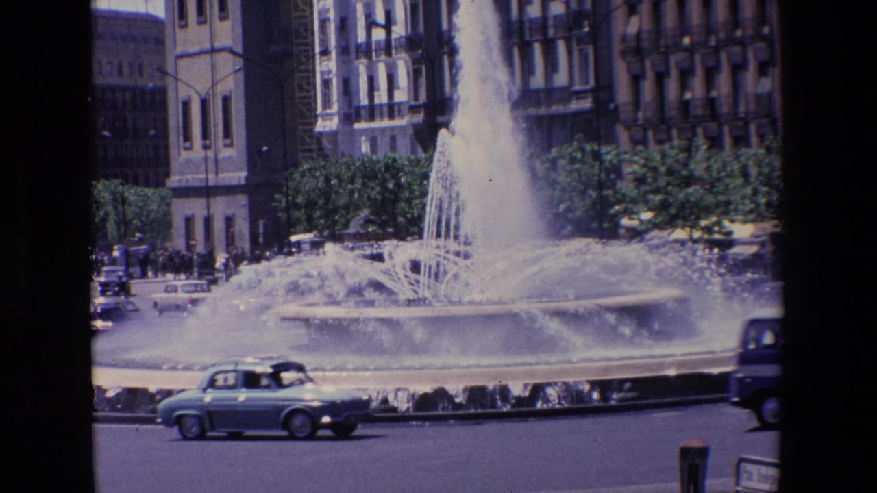 1967 Madrid home-movie frame showing a large public fountain, passing compact cars, distant pedestrians, trees, and multi-story buildings beyond the street.