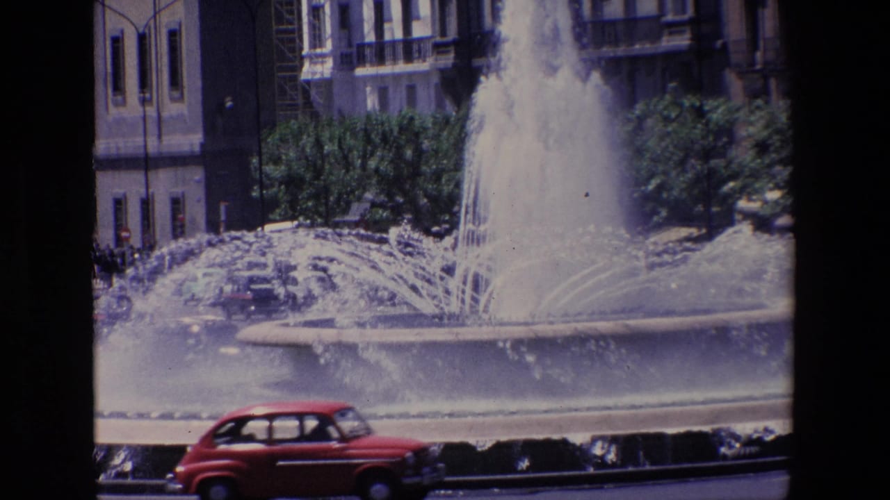 Red compact car passes in front of a large spraying fountain and city buildings.