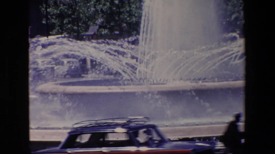 1967 Madrid home-movie frame showing fountain jets and mist near the street, with a dark car or wagon partially visible at the bottom of the frame.