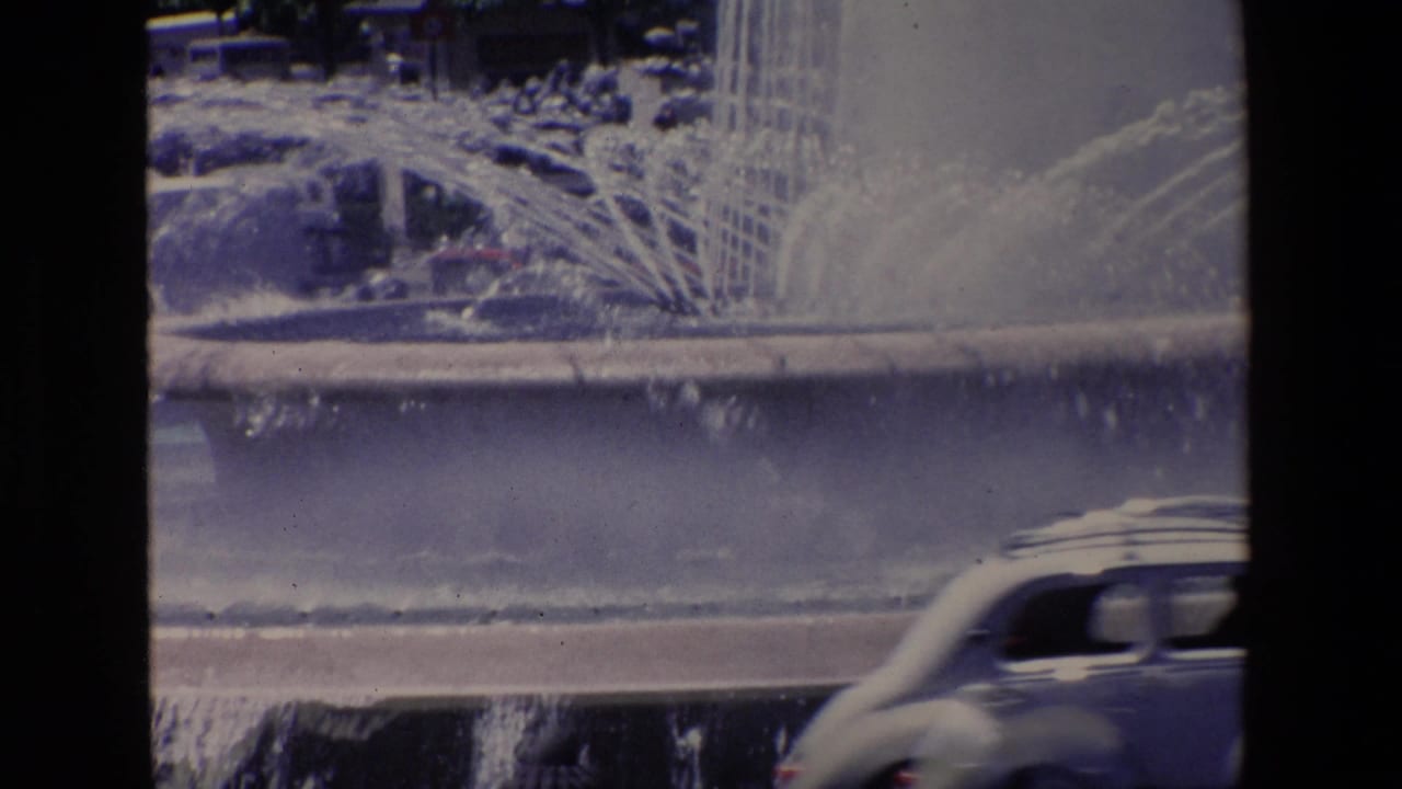 1967 Madrid home-movie frame showing a public fountain spraying water toward a road while a rounded car passes along the wet street.
